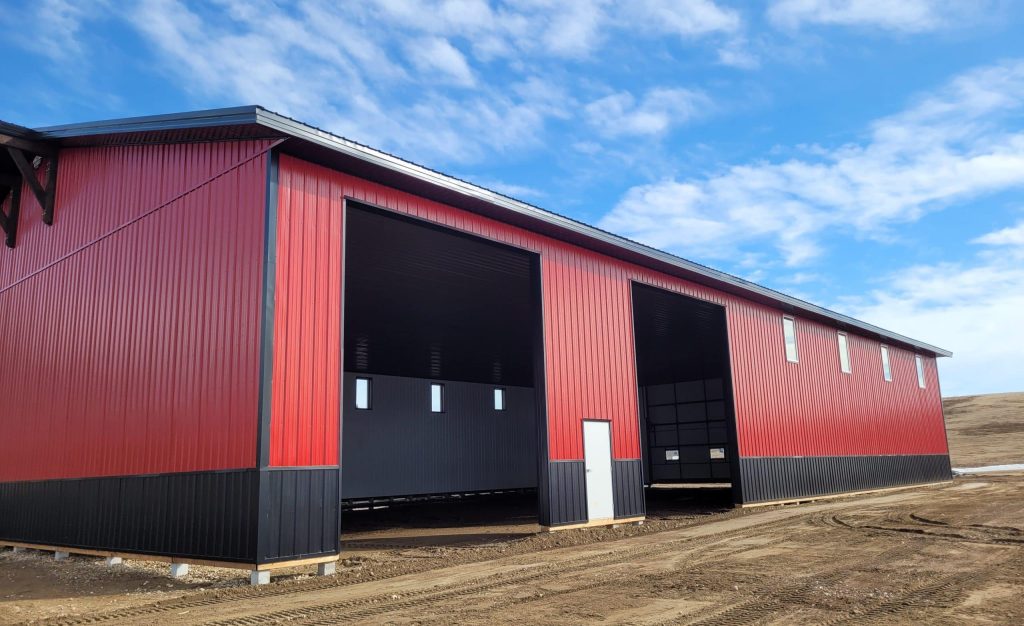 A dark red agricultural post-frame shop with two overhead doors, timber accents, crows peak, and a bi-fold door