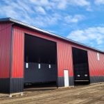 A dark red agricultural post-frame shop with two overhead doors, timber accents, crows peak, and a bi-fold door