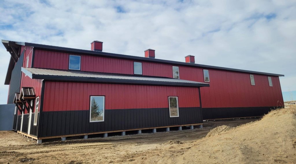 A dark red agricultural post-frame shop with two overhead doors, timber accents, crows peak, and a bi-fold door