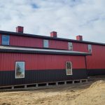 A dark red agricultural post-frame shop with two overhead doors, timber accents, crows peak, and a bi-fold door