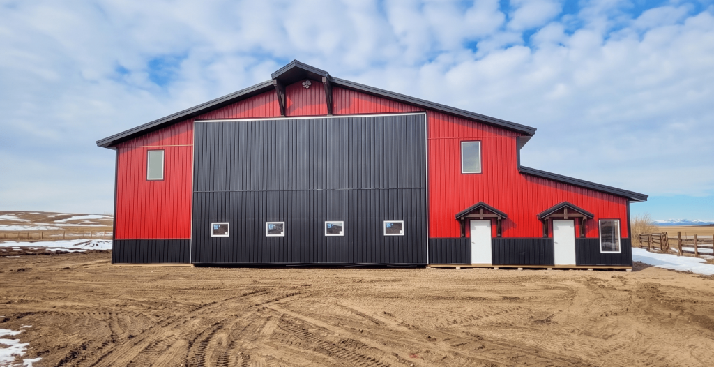 A dark red agricultural post-frame shop with two overhead doors, timber accents, crows peak, and a bi-fold door