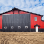 A dark red agricultural post-frame shop with two overhead doors, timber accents, crows peak, and a bi-fold door