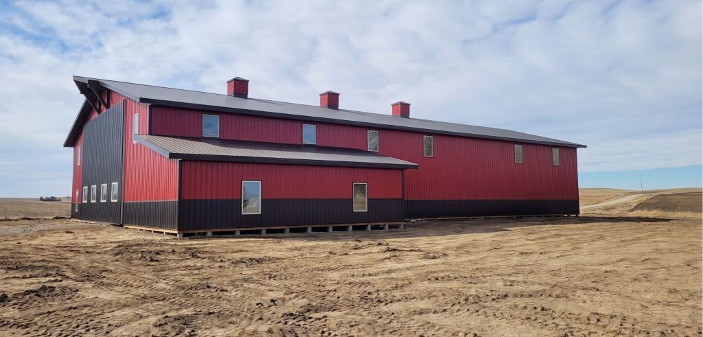 A dark red agricultural post-frame shop with two overhead doors, timber accents, crows peak, and a bi-fold door