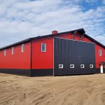 A dark red agricultural post-frame shop with two overhead doors, timber accents, crows peak, and a bi-fold door