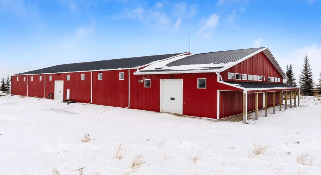 A dark red post-frame riding arena with soffits, sliding doors, and two overhead doors