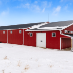 A dark red post-frame riding arena with soffits, sliding doors, and two overhead doors