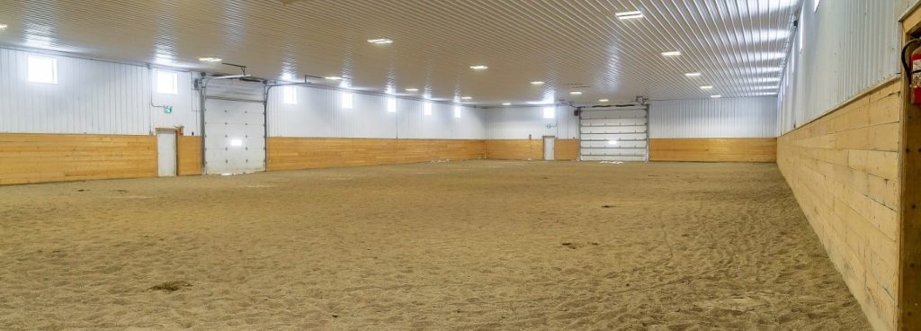 A dark red post-frame riding arena with soffits, sliding doors, and two overhead doors