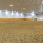 A dark red post-frame riding arena with soffits, sliding doors, and two overhead doors