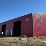 A bright red post-frame equestrian barn with overhead doors and a black roof