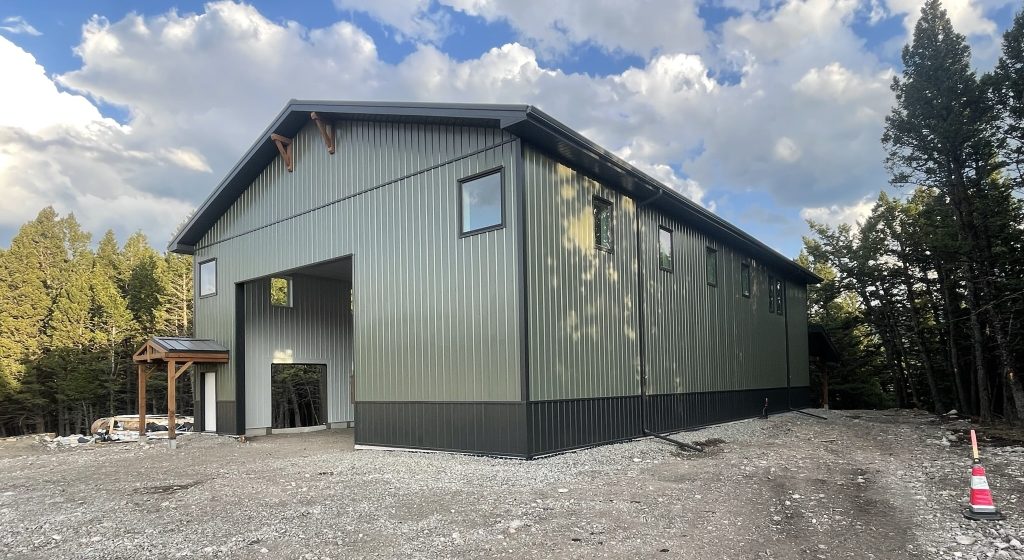 A dark sage post-frame shouse with open lean-tos, overhead doors, soffits, wainscoting, amd timber accents