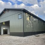 A dark sage post-frame shouse with open lean-tos, overhead doors, soffits, wainscoting, amd timber accents