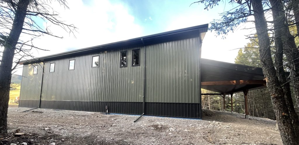 A dark sage post-frame shouse with open lean-tos, overhead doors, soffits, wainscoting, amd timber accents