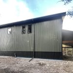 A dark sage post-frame shouse with open lean-tos, overhead doors, soffits, wainscoting, amd timber accents