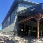 A dark sage post-frame shouse with open lean-tos, overhead doors, soffits, wainscoting, amd timber accents