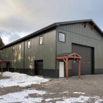 A dark sage post-frame shouse with open lean-tos, overhead doors, soffits, wainscoting, and timber accents