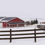 A dark red post-frame riding arena with soffits, sliding doors, and two overhead doors