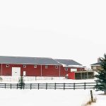 A dark red post-frame riding arena with soffits, sliding doors, and two overhead doors