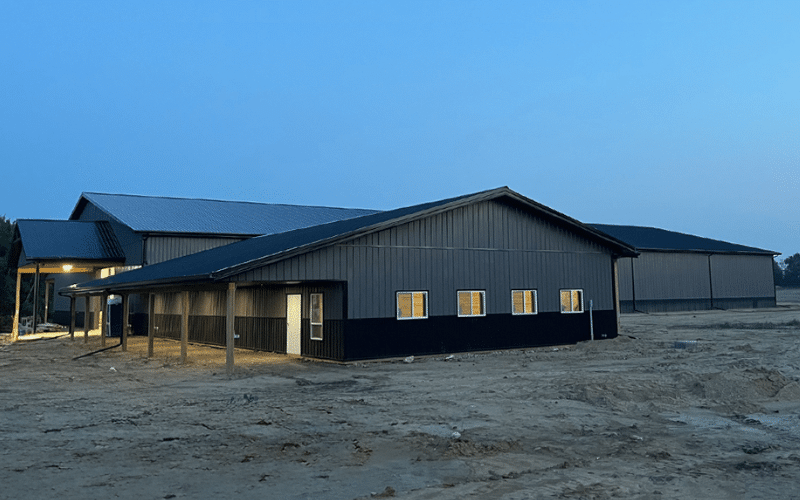A charcoal post-frame riding arena with overhead doors, crows peak, wainscoting, and soffits