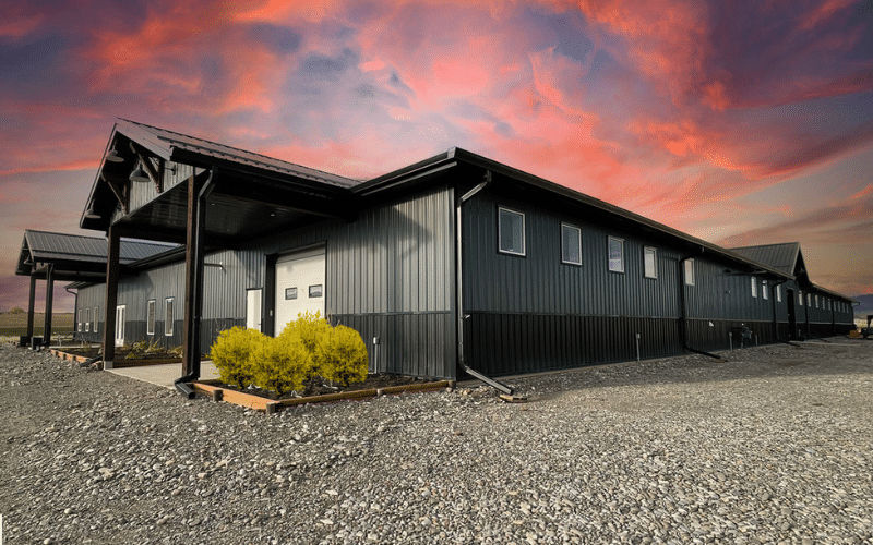 A gunmetal post-frame riding arena with wainscoting, overhead doors, soffits, and timber accents
