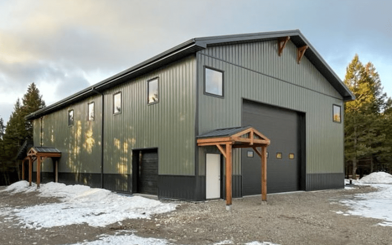 A dark sage post-frame shouse with open lean-tos, overhead doors, soffits, wainscoting, amd timber accents