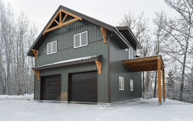 A dark sage custom post-frame residential build with overhead doors, awnings, timber accents, and soffits