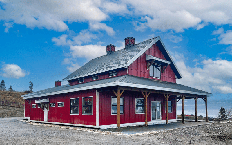 A dark red post-frame barndominium with cupolas, attached garage with overhead doors, open lean-tos, soffits, and timber accents