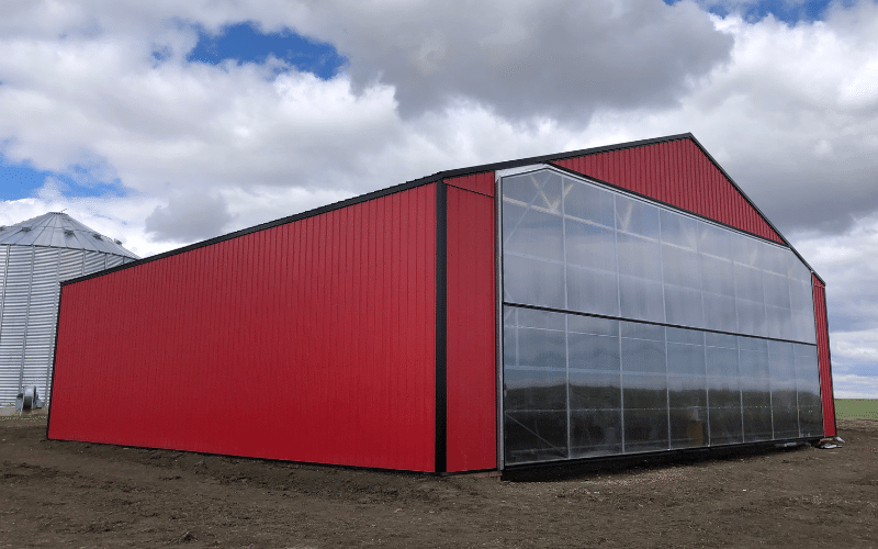 A bright red post-frame agricultural storage building with a clear carbonate bi-fold door