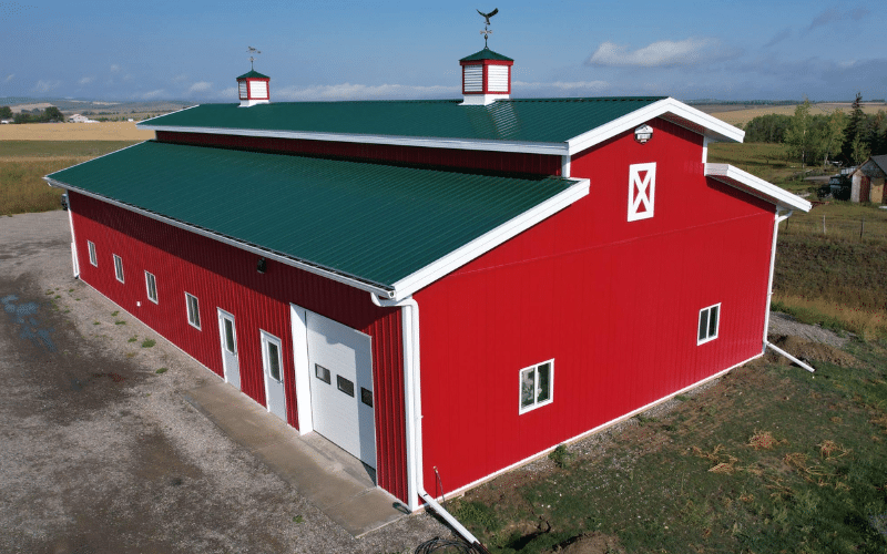 A dark red post-frame equestrian barn with a melchers green roof, soffits, and overhead door