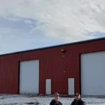 A bright red post-frame equestrian barn with overhead doors and a black roof