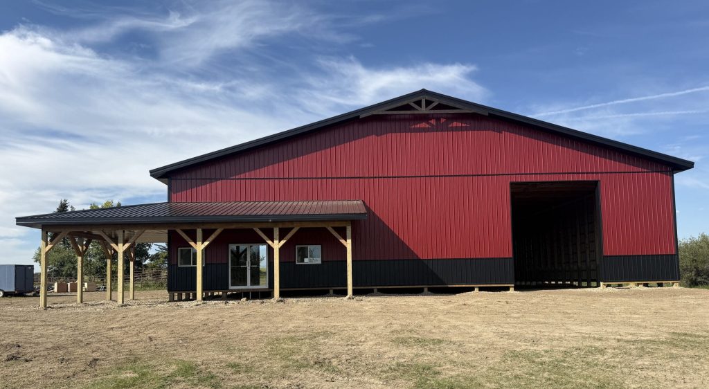 A dark red post-frame equestrian barn with soffits, overhead doors, a wrap around porch, and timber accents