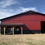 A dark red post-frame equestrian barn with soffits, overhead doors, a wrap around porch, and timber accents