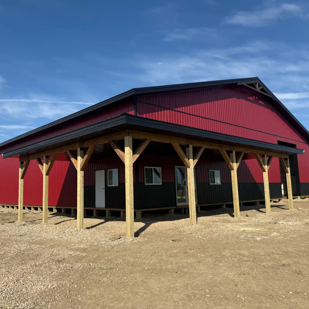 A dark red post-frame equestrian barn with soffits, overhead doors, a wrap around porch, and timber accents