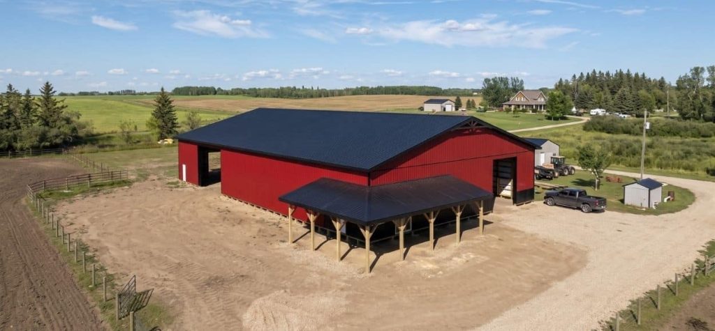 A dark red post-frame equestrian barn with soffits, overhead doors, a wrap around porch, and timber accents