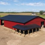 A dark red post-frame equestrian barn with soffits, overhead doors, a wrap around porch, and timber accents