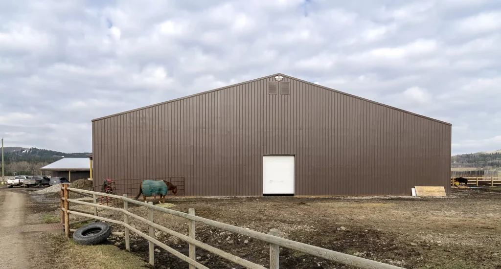 A coffee brown post-frame equestrian riding arena with overhead doors and an open lean-to