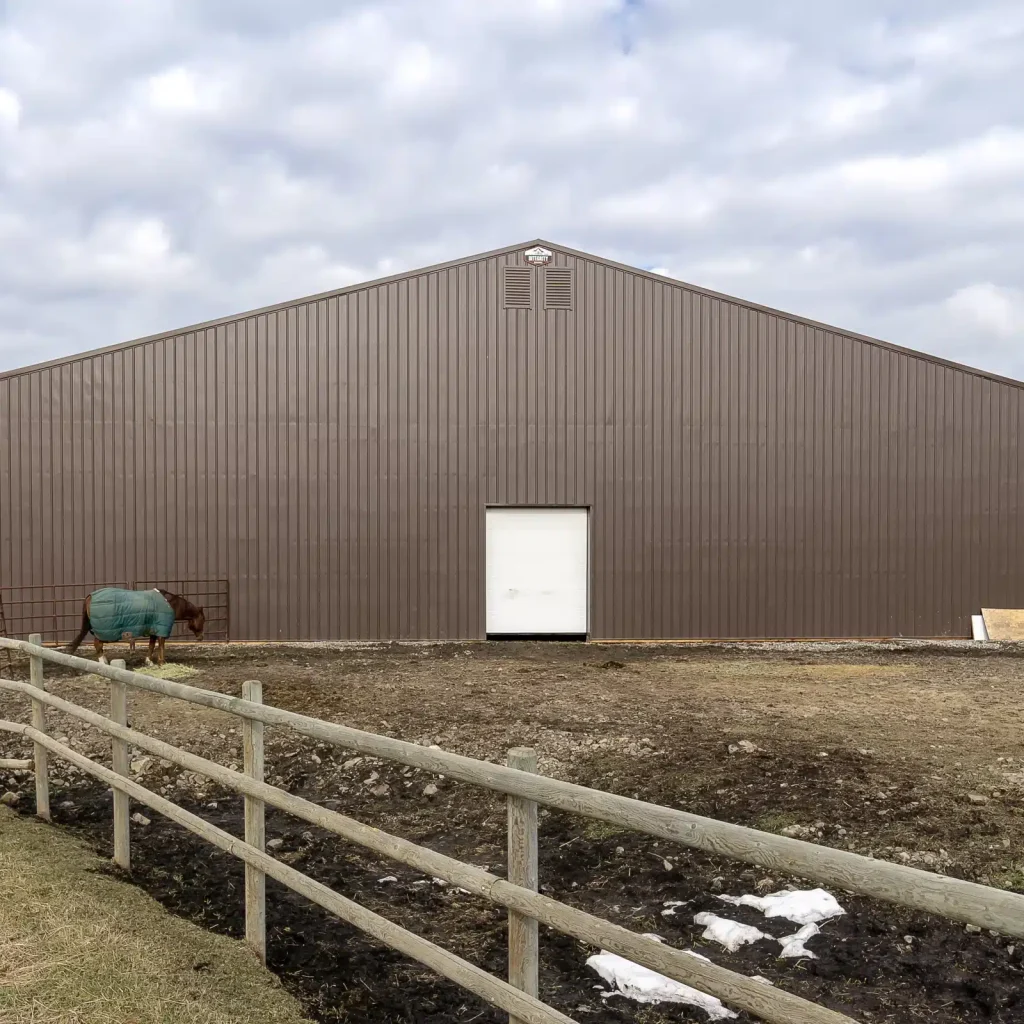 A coffee brown post-frame equestrian riding arena with overhead doors and an open lean-to