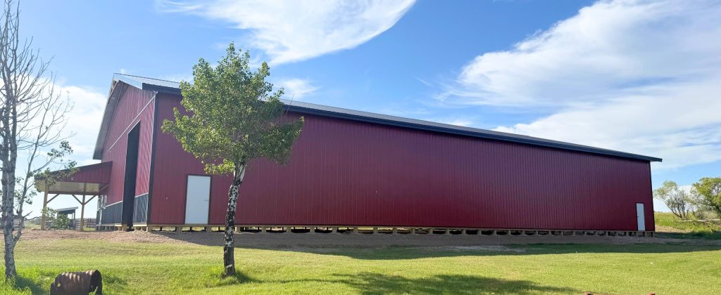 A dark red post-frame equestrian barn with soffits, overhead doors, a wrap around porch, and timber accents