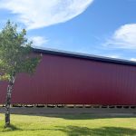 A dark red post-frame equestrian barn with soffits, overhead doors, a wrap around porch, and timber accents
