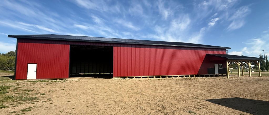A dark red post-frame equestrian barn with soffits, overhead doors, a wrap around porch, and timber accents