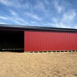 A dark red post-frame equestrian barn with soffits, overhead doors, a wrap around porch, and timber accents