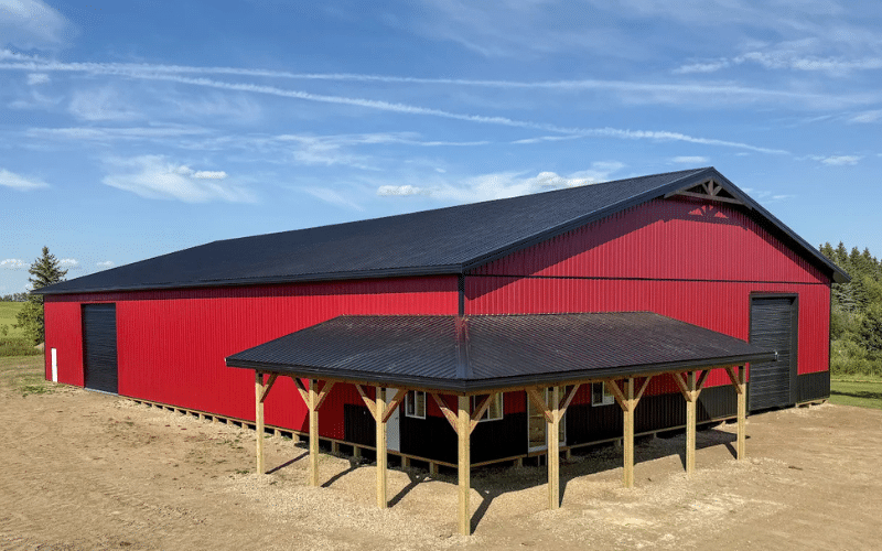A dark red post-frame equestrian barn with soffits, overhead doors, a wrap around porch, and timber accents