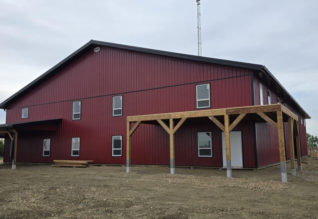 A dark red post-frame equestrian barn and office with open lean-to and deck