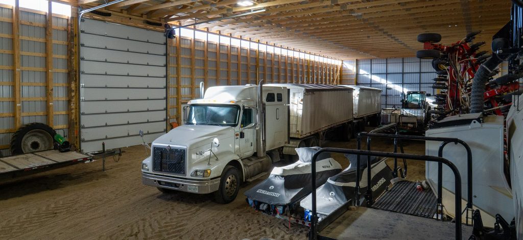 A dark red post-frame machine shed with a bi-fold door, wall lights, and overhead doors