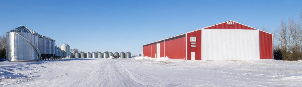 A dark red post-frame machine shed with a bi-fold door, wall lights, and overhead doors