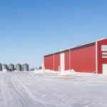 A dark red post-frame machine shed with a bi-fold door, wall lights, and overhead doors