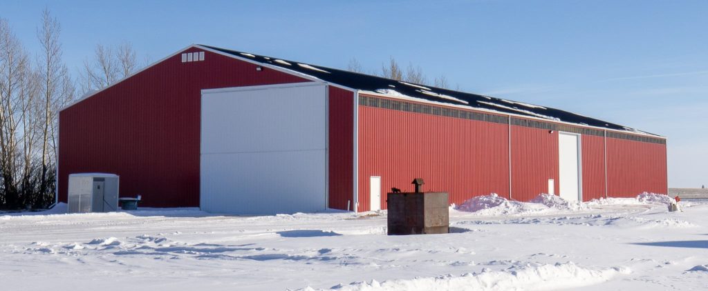 A dark red post-frame machine shed with a bi-fold door, wall lights, and overhead doors