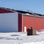 A dark red post-frame machine shed with a bi-fold door, wall lights, and overhead doors