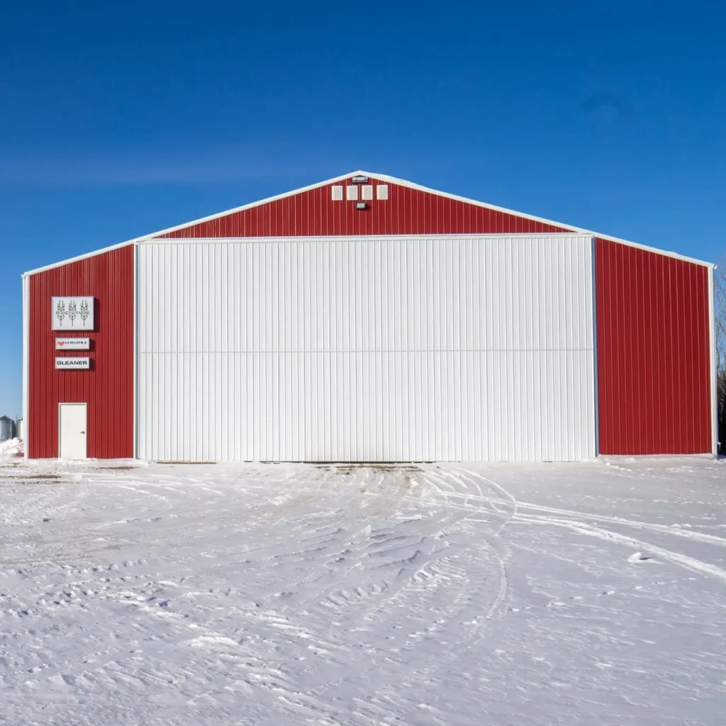 A dark red post-frame machine shed with a bi-fold door, wall lights, and overhead doors