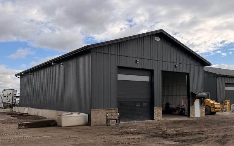 A gunmetal post-frame storage building with wainscoting, soffits, and overhead doors